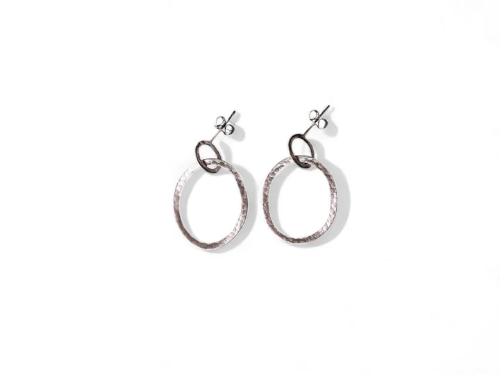 A pair of elegant sterling silver earrings with a hammered texture. Each hand-forged earring features a larger circular hoop connected to a smaller loop that attaches to a simple stud fastening. The Carran Drops by Lynsey de Burca are placed on a white background.