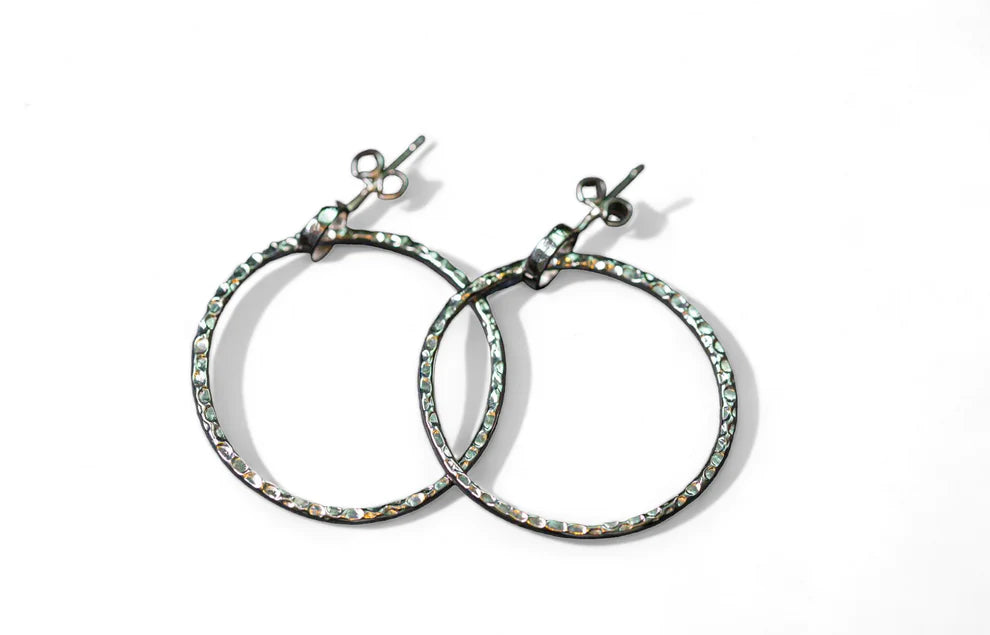 A pair of Carran Hoops from Lynsey de Burca, laid out against a white background. Each earring has a post and butterfly clasp for fastening. The textured hoop earrings intersect at one point, resembling organic links.