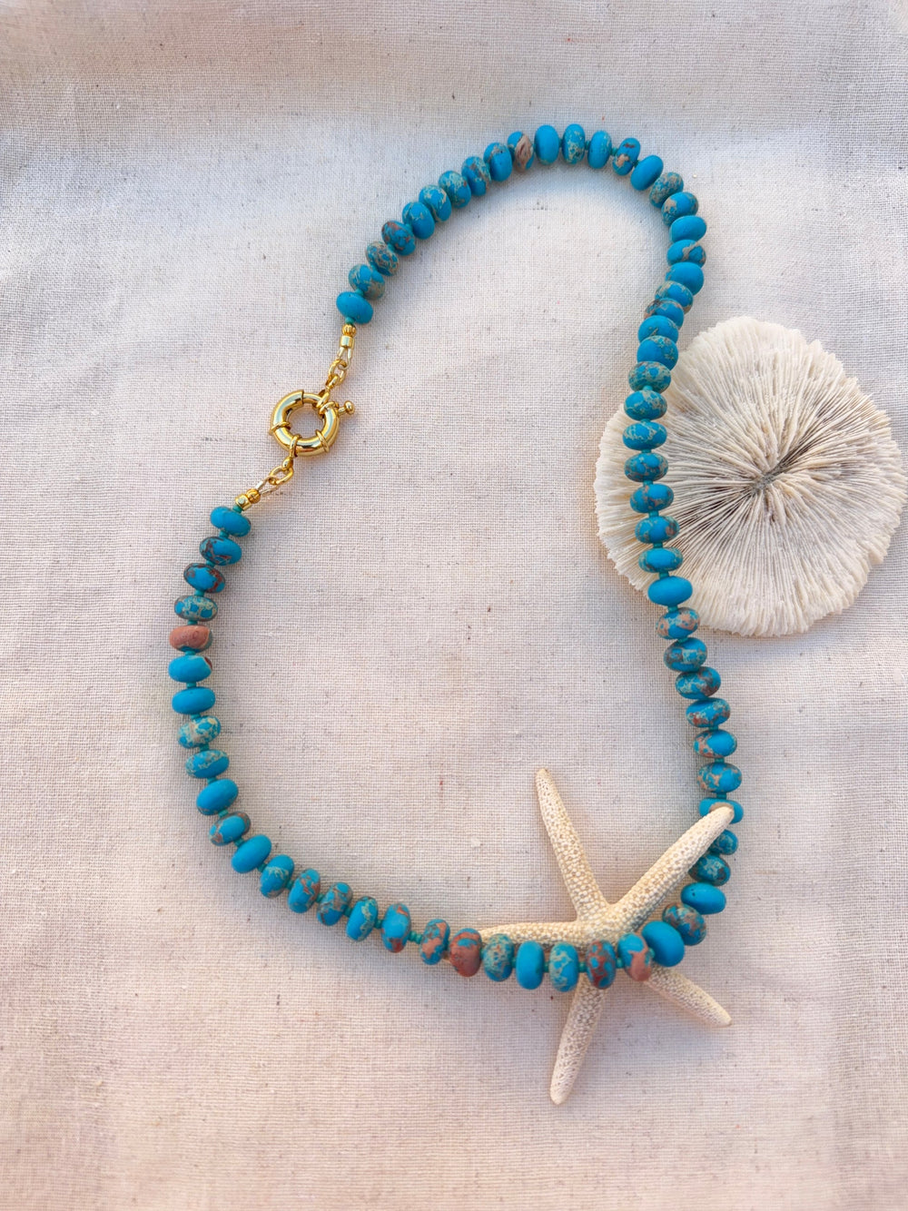 The Turquoise Candy Bead Necklace by Kybalion Jewellery features turquoise beads and a gold-plated clasp, displayed in a loose loop on light fabric with a starfish and seashell accenting this handmade piece.