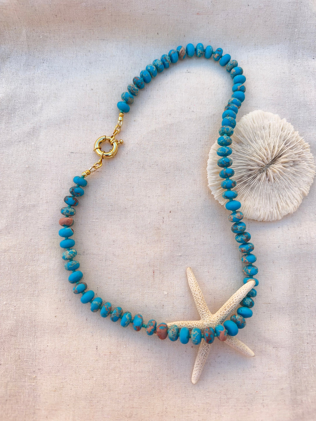 The Turquoise Candy Bead Necklace by Kybalion Jewellery features turquoise beads and a gold-plated clasp, displayed in a loose loop on light fabric with a starfish and seashell accenting this handmade piece.