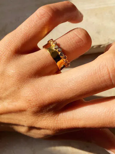 A close-up of a hand wearing two gold rings on the index finger. One is a solid, chunky Clarence Ring by Shyla, and the other is decorated with small, evenly spaced diamonds. The hand is positioned in natural lighting, casting soft shadows.