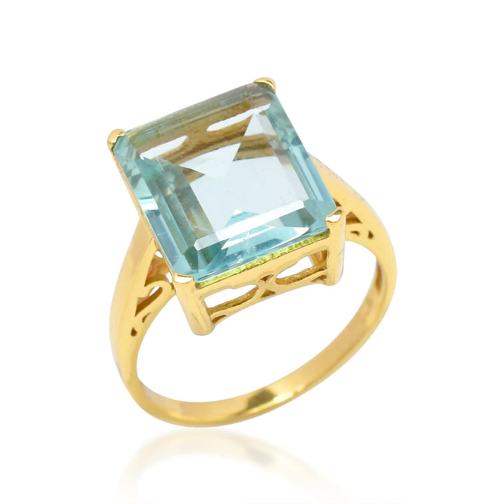 The Claudia Ring by Shyla is a vintage inspired ring crafted from 22 carat gold, featuring a large, rectangular light blue aquamarine gemstone in a prong setting. The ring band has decorative cut-out designs on the sides, adding a touch of elegance to the overall design.