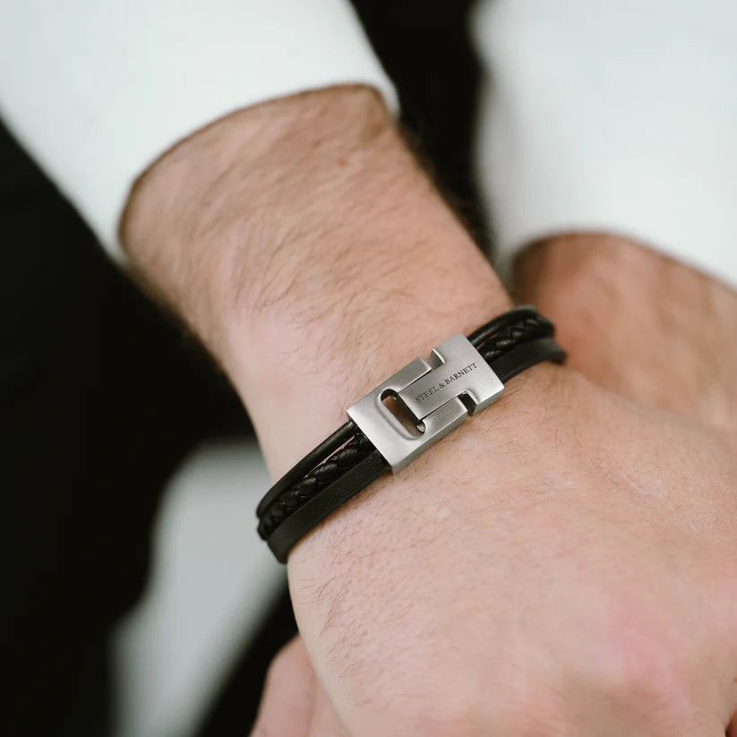 Close-up of a person wearing a stylish Cole - Black bracelet by Steel and Barnett on their wrist. The bracelet features a sleek stainless steel clasp with the engraved text "STEEL & BARNETT" and consists of multiple black braided genuine leather bands. The person is dressed in a white shirt and dark jacket.