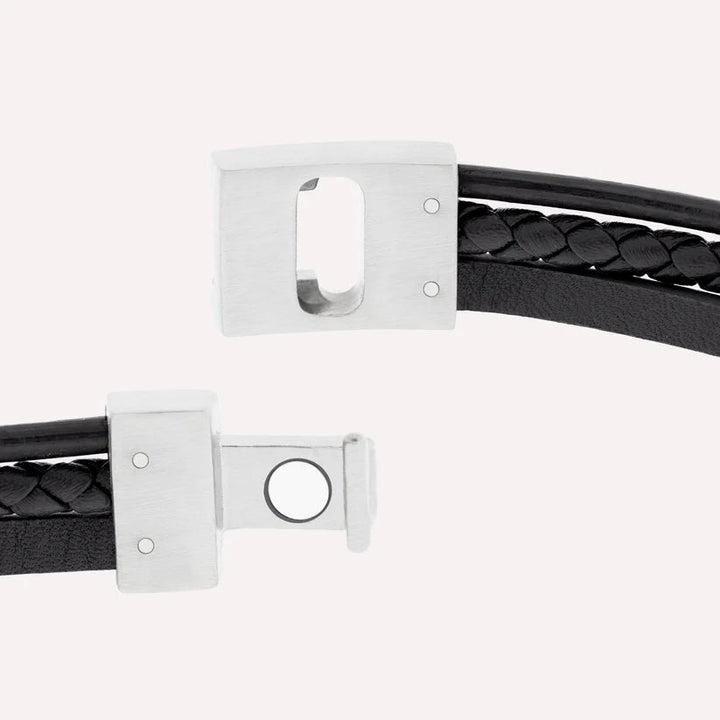 A close-up of a Steel and Barnett Cole - Black bracelet made from genuine black leather, featuring a braided design along the center. The stainless steel clasp is partially open, revealing the locking mechanism. The background is a plain, light color.