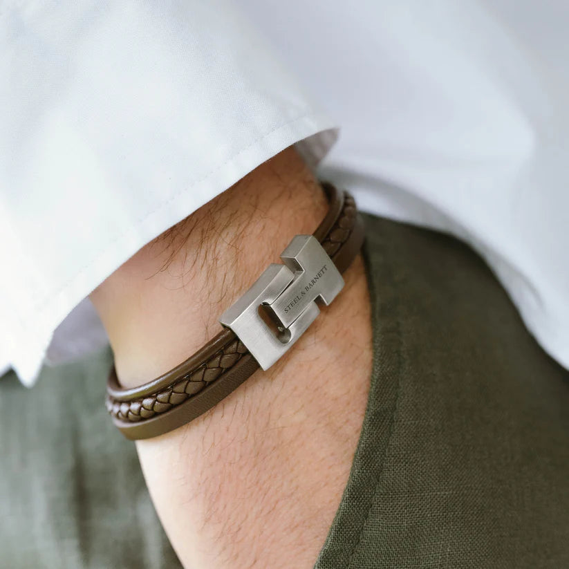 Close-up of a person's wrist adorned with the Cole - Brown bracelet by Steel and Barnett, featuring genuine leather cords and a stainless steel clasp engraved with the initials "GH." The person is dressed in a white shirt and green pants.