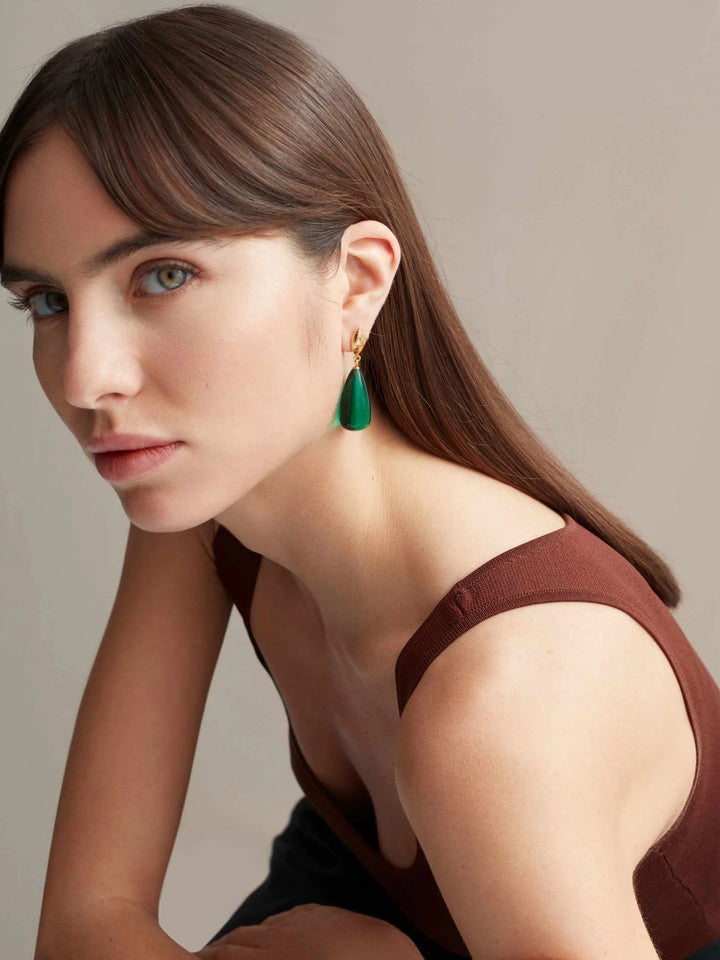 A woman with straight brown hair and bangs, wearing a sleeveless brown top, looks into the camera. She has green eyes and is adorned with a single Shyla Corine Earring in green, featuring a golden accent. The background is neutral.
