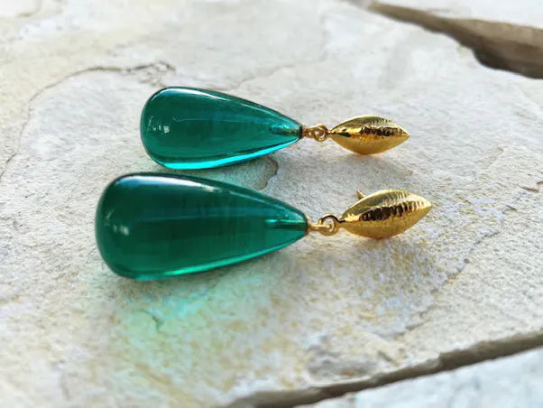 A pair of Corine Earrings by Shyla in elegant, hypoallergenic teardrop-shaped emerald green gemstones set in gold-plated mounts. Lying on a textured, light-colored stone surface, the smooth and polished green gems contrast beautifully with the matte gold settings.