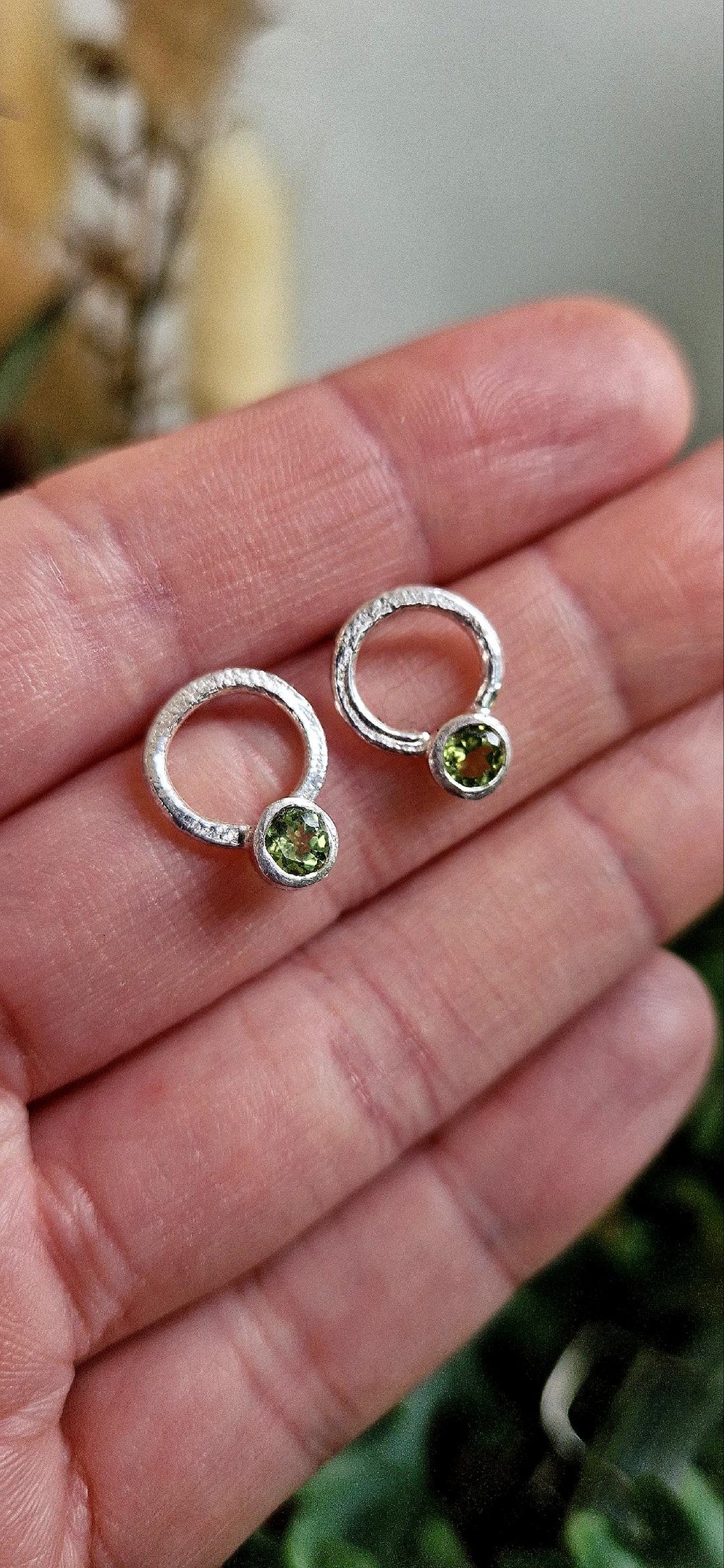 A close-up shows a hand holding Caroline Stokesberry-lee's Circle of Light Earrings in Peridot—small silver hoops each with a round green gemstone at the base. The softly blurred background highlights the elegance of these unique earrings.