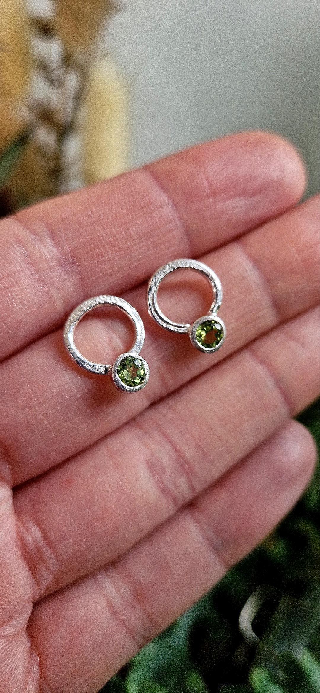 A close-up shows a hand holding Caroline Stokesberry-lee's Circle of Light Earrings in Peridot—small silver hoops each with a round green gemstone at the base. The softly blurred background highlights the elegance of these unique earrings.
