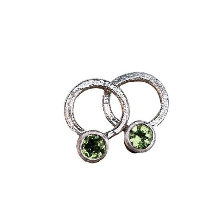 The Circle of Light Earrings in Peridot by Caroline Stokesberry-lee are silver circular studs featuring a small round green peridot at the base, offering a timeless style for gemstone earrings.