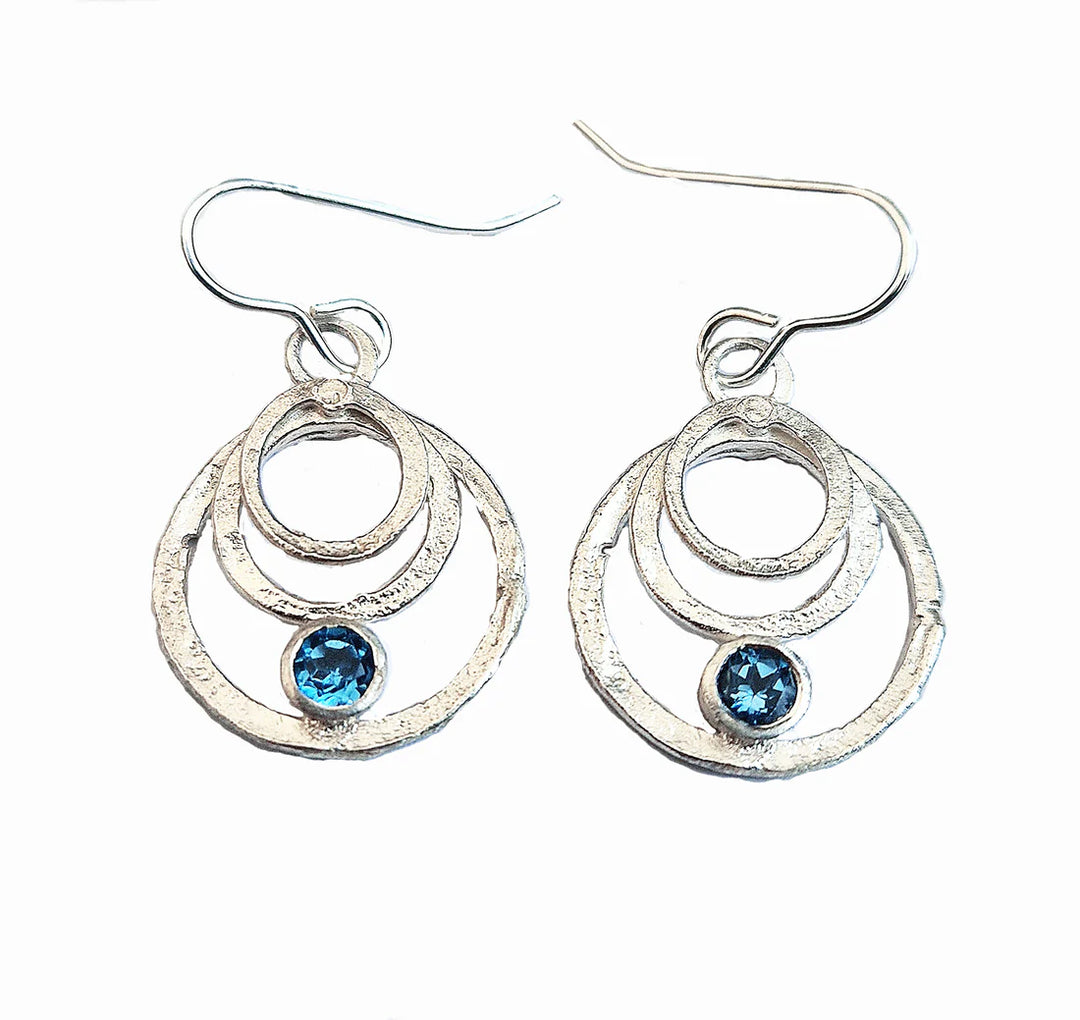 The Caroline Stokesberry-lee Rays of Light Earrings in Blue Topaz feature sterling silver with three textured concentric circles and a blue topaz gemstone, dangling elegantly from classic hook-style ear wires.
