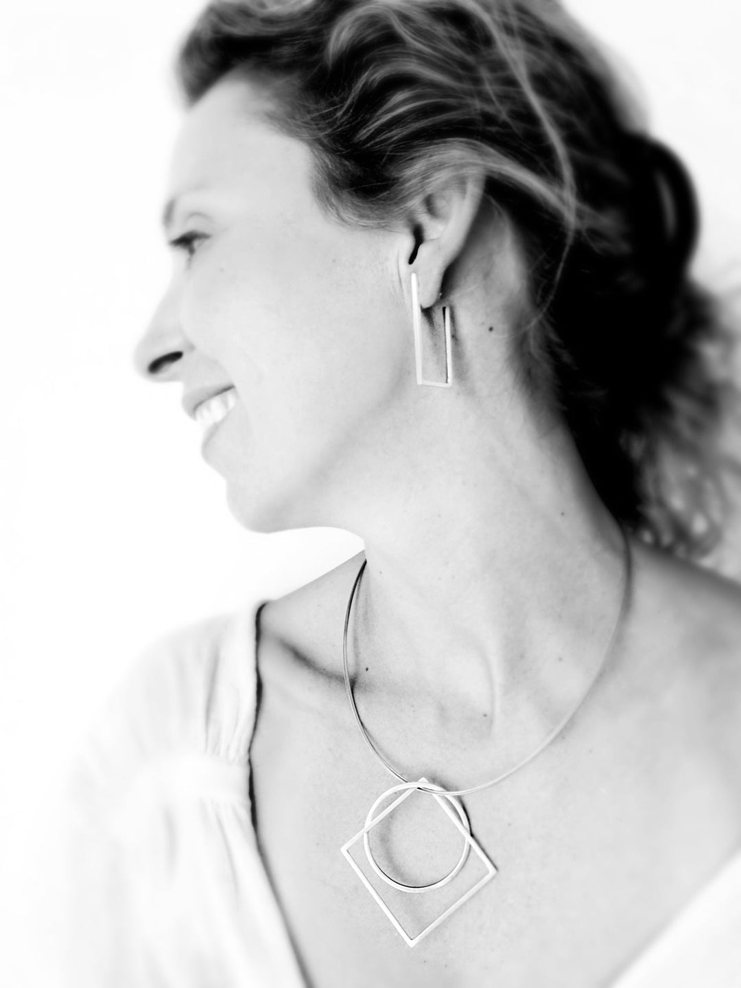 A black-and-white photo of a person smiling and looking to the side. The individual is wearing handmade jewelry from MarCucurella's Cub collection, including a necklace with overlapping squares and circles, and matching Cub Long Silver Earrings. The photo has a soft, blurred background.