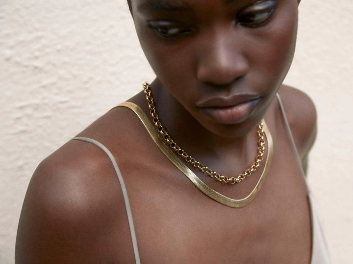 A close-up of a woman wearing a sleeveless top, looking down and away from the camera. She is adorned with two elegant necklaces: a Shyla Curzon Gold Chain Necklace and a thinner, braided chain. The background is a textured, off-white wall.