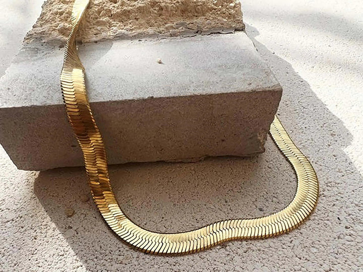 A chunky 22 carat gold serpentine Curzon Gold Chain Necklace by Shyla is elegantly draped over a rough-edged concrete block on a textured beige surface, with sunlight casting soft shadows across the scene.