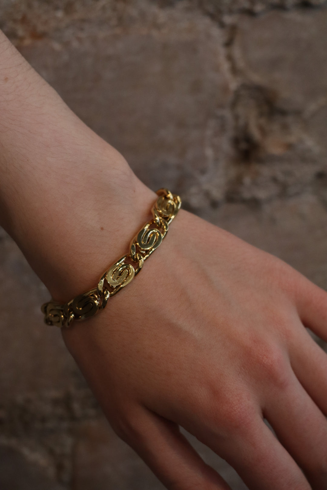 A close-up of a wrist adorned with the Zubeldia Caesar Chain Gold Plated Scroll Chunky Bracelet, featuring intricate oval-shaped links, set against a textured beige stone background.