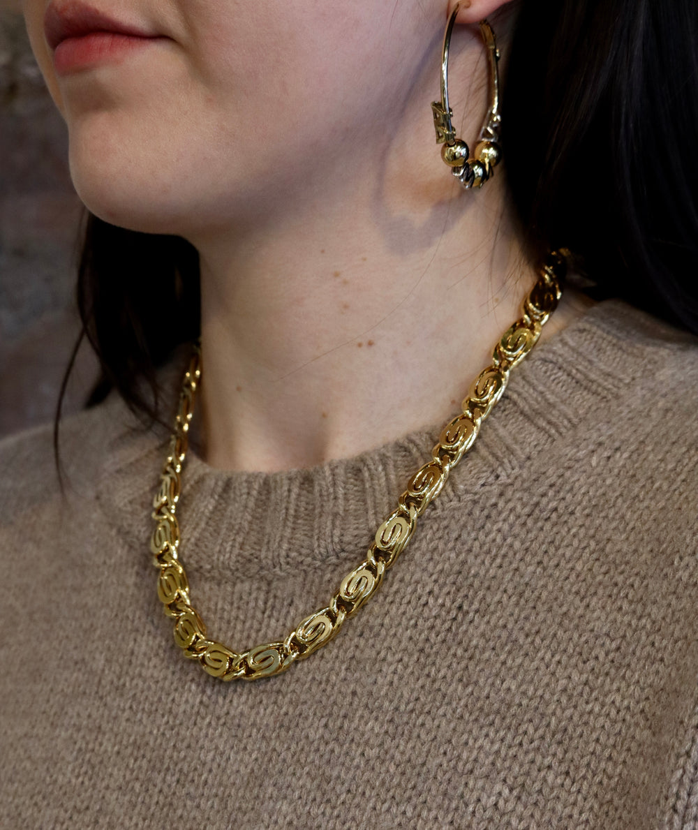 A person wearing the Zubeldia Caesar Chain Gold Scroll Chunky Necklace and large gold hoop earrings, paired with a beige knit sweater. The image is cropped to show only the lower face, neck, and upper chest.