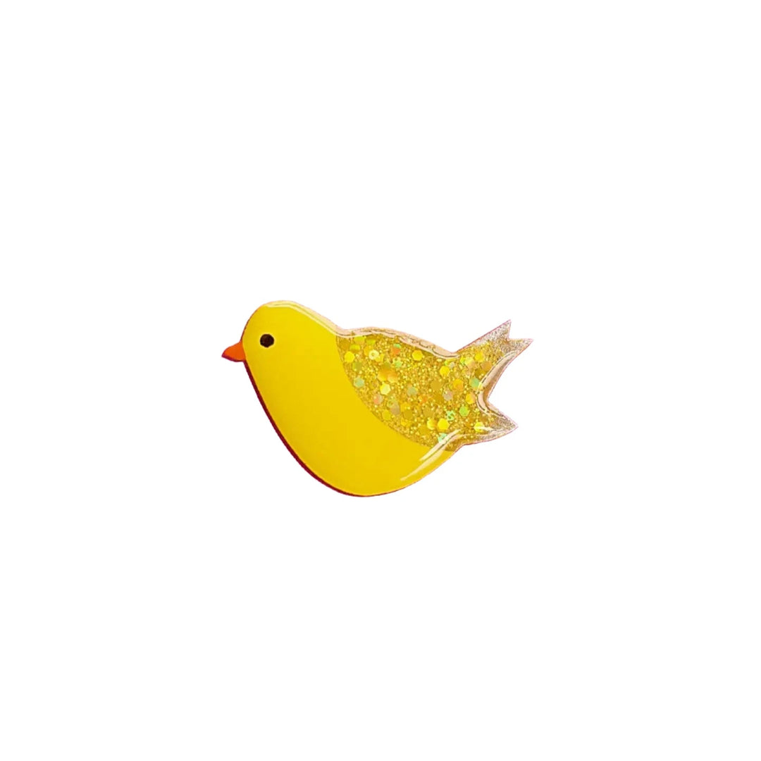 A small, yellow bird-shaped pin with a red beak. The bird's wing and tail, now encased in a resin coating, are filled with glitter for a sparkly appearance. This whimsical design is simple yet enchanting and is set against a white background. Introducing the Canary Pin Brooch by Naoi.