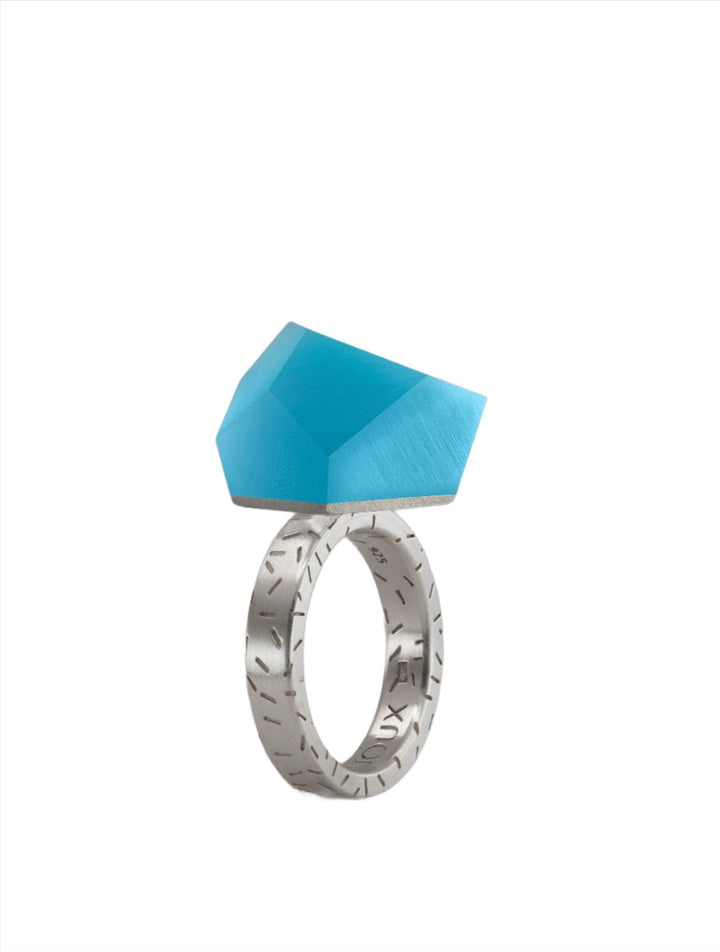 A sterling silver ring with engraved patterns on the band featuring a large, faceted, transparent blue gemstone. The design is modern and geometric, with clean lines and a bold aesthetic. The Fruit Bijoux Ring Top VU Crystals Capri Blue by Fruit Bijoux is displayed against a plain white background.