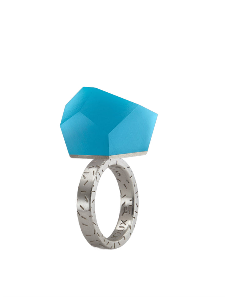 A modern Fruit Bijoux Ring Top VU Crystals Capri Blue with a large geometric turquoise stone on top of a Sterling silver band. The band is engraved with abstract patterns, adding a unique touch to the contemporary design. The faceted stone brings a bold and artistic look to the piece.