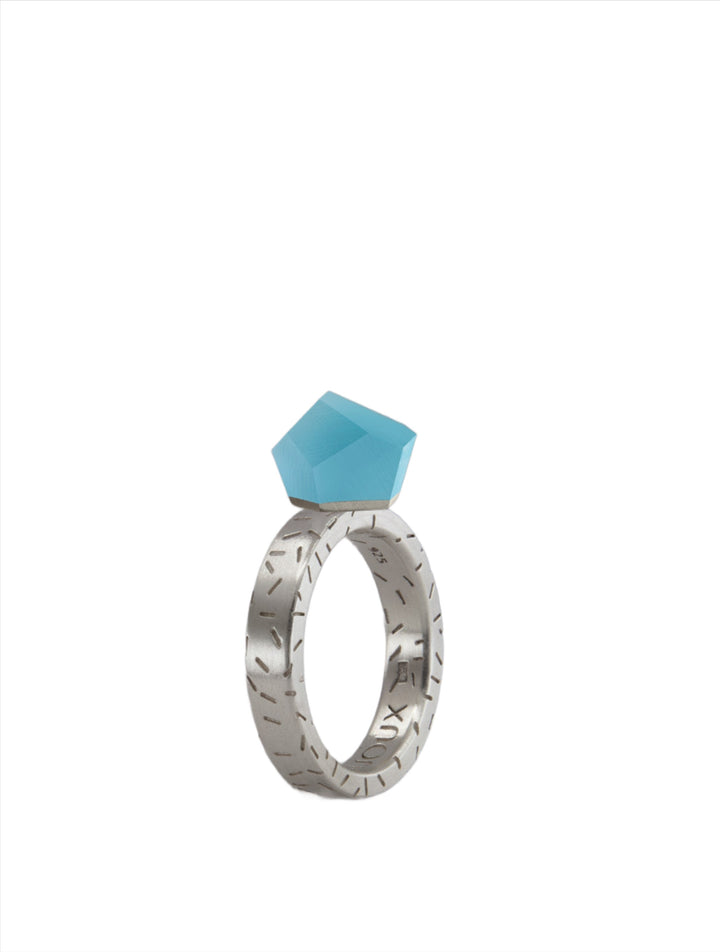 A Fruit Bijoux Fruit Bijoux Ring Top VU Crystals Capri Blue with a geometric engraving pattern, featuring a large, faceted, sky-blue gemstone positioned at the top. The background is plain white, highlighting the ring's design and the vibrant color of the gemstone.