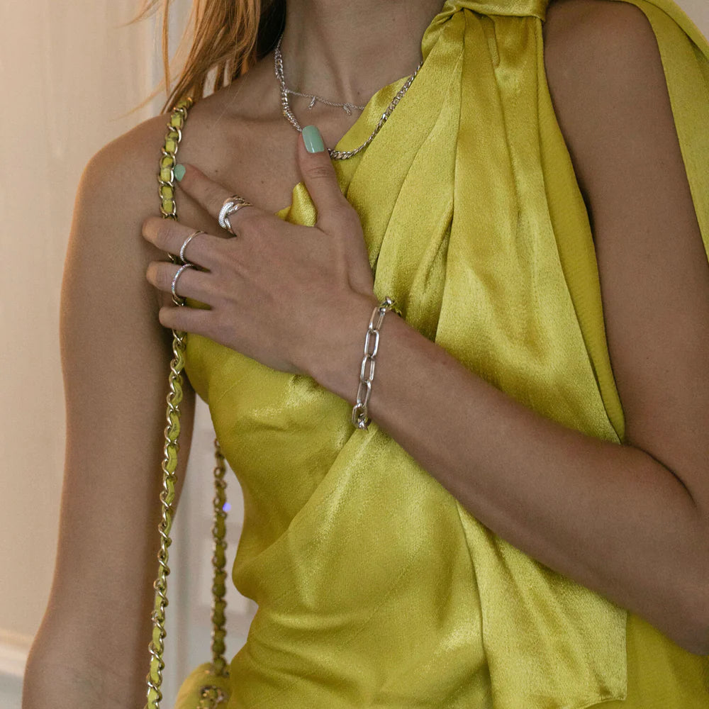 A woman wearing a shiny yellow sleeveless top holds her hand to her chest, showcasing silver rings, a Sif Jacobs Capri Bracelet Silver, a necklace, and a matching yellow shoulder bag with a chain strap. Her nails are light green.