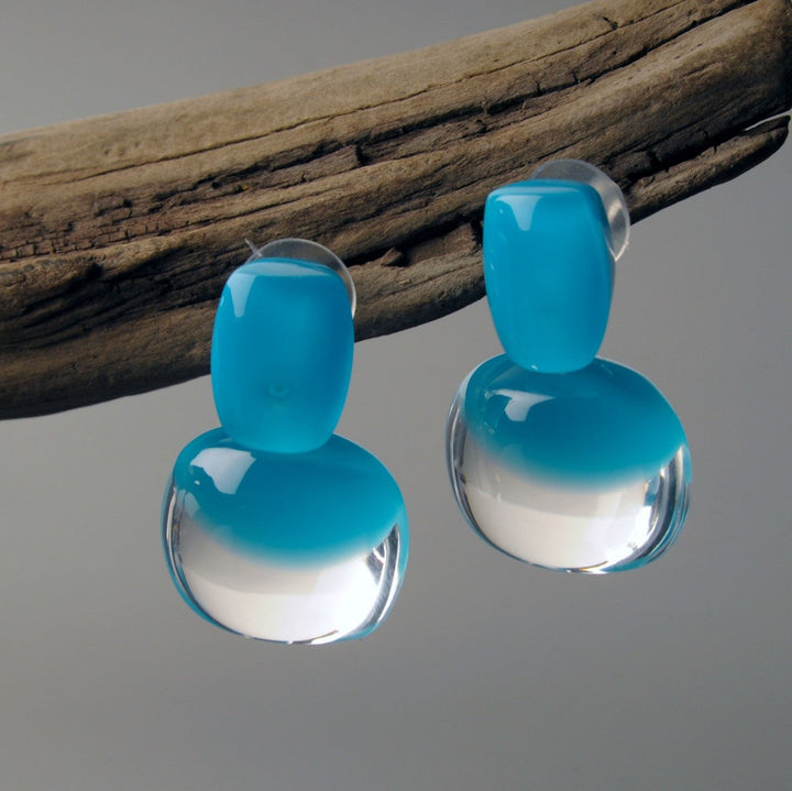 A pair of MX.design Acrylic Capri Blue Earrings with transparent lower halves hang from a rustic piece of wood. The earrings have a smooth, glossy finish and reflect light, displaying a gradient from solid blue at the top to clear at the bottom.