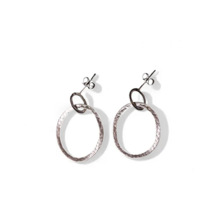 A pair of elegant sterling silver earrings with a hammered texture. Each hand-forged earring features a larger circular hoop connected to a smaller loop that attaches to a simple stud fastening. The Carran Drops by Lynsey de Burca are placed on a white background.