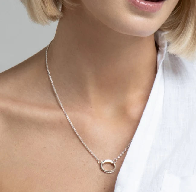 A person wearing a delicate silver chain necklace with a Carran Mix Pendant by Lynsey de Burca and matching Carran earrings. They are dressed in a white, off-the-shoulder top, and their short blonde hair is partially visible. The background is plain and light-colored, focusing attention on the jewelry and clothing.