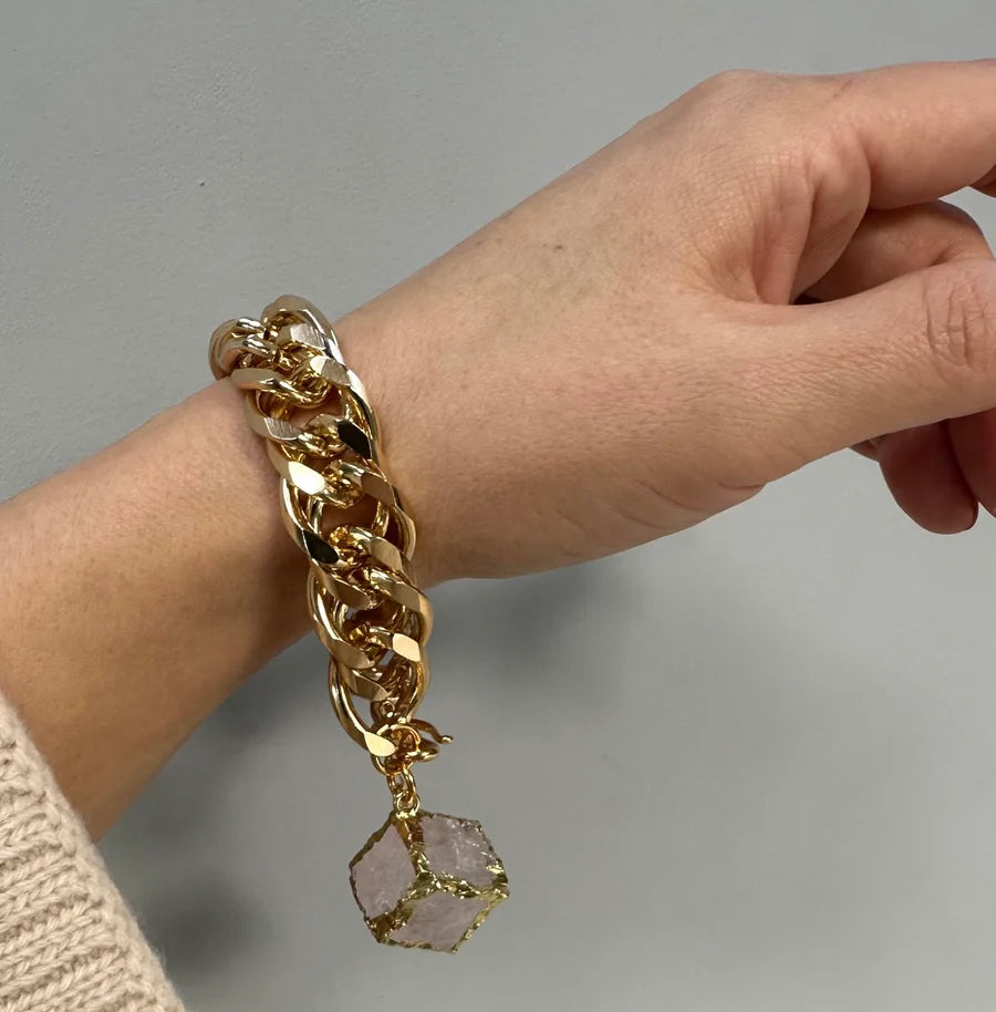 A hand wears the Aria V Carrie Bracelet—a chunky gold-plated piece with a large, cube-shaped stone pendant wrapped in gold wire—handmade in Dublin, shown against a plain light gray background.