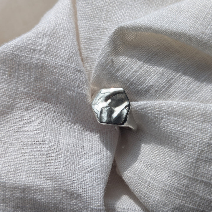 The Causeway Coastal Silver Signet Ring by Becca Calmont, crafted from recycled sterling silver with an organic top inspired by the Giant’s Causeway, rests on textured white fabric in natural light.