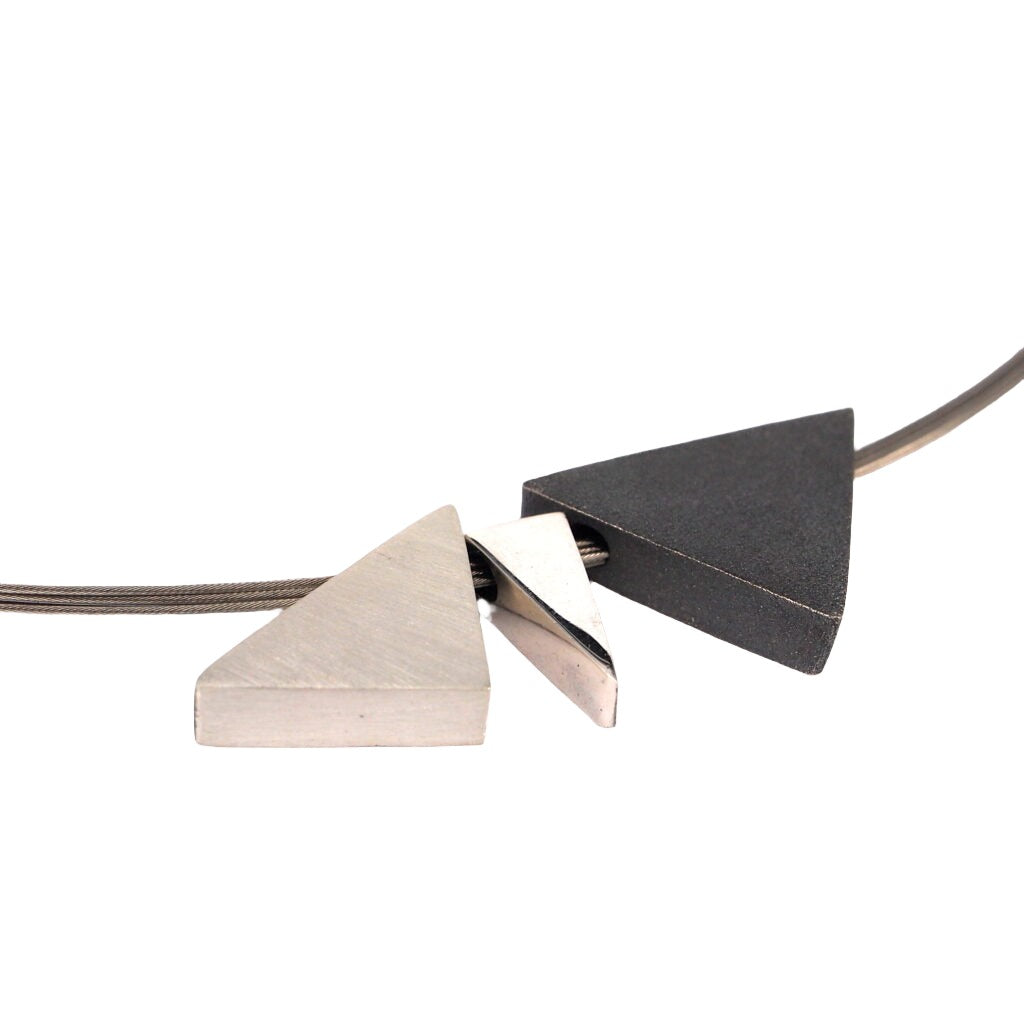 A modern necklace featuring intertwined triangular pendants on a sleek metallic wire. One triangle is sterling silver, another is dark matte oxidized silver, and the third is positioned between them, creating a geometric and minimalist design in this exquisite piece of handmade jewelry. Introducing the Keops 3 Part Silver Pendant by MarCucurella.