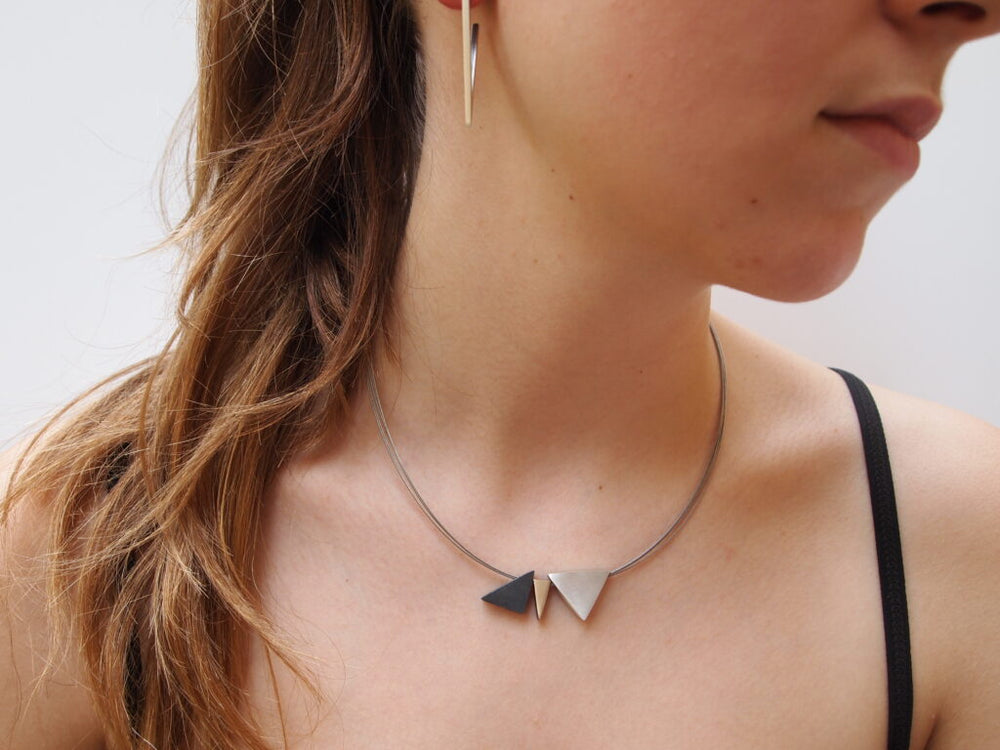 Close-up of a person wearing a minimalist silver necklace with the Keops 3 Part Silver & Gold Pendant from MarCucurella and a thin, modern silver earring. The person has long hair and is wearing a black strap top, facing slightly away from the camera. The pieces are handmade jewelry from a Barcelona studio.