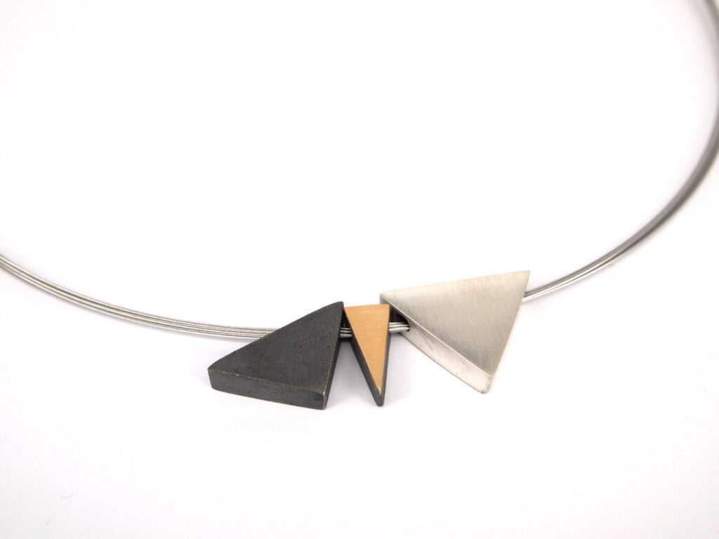 A minimalistic Keops 3 Part Silver & Gold Pendant with three geometric pendants on a thin, stainless steel wire. Handmade jewelry from the MarCucurella studio in Barcelona, the pendants include a black metal triangle, a small 18ct matt gold triangle, and a larger silver triangle, positioned next to each other. The background is white.