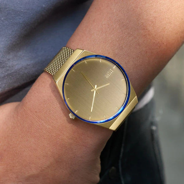 A Storm Cirero Gold Watch with a minimalist, contemporary design and a mesh strap graces a person's left wrist. The watch, crafted by Storm Watches, features a blue accent around the edge of the face and three hands for hours, minutes, and seconds. The background is slightly blurred.