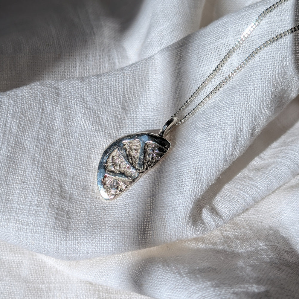 The Becca Calmont Citrus Slice Pendant, a silver necklace with a fruit-slice design and diamond cut curb chain, rests on soft white fabric in natural sunlight, its details shimmering subtly.