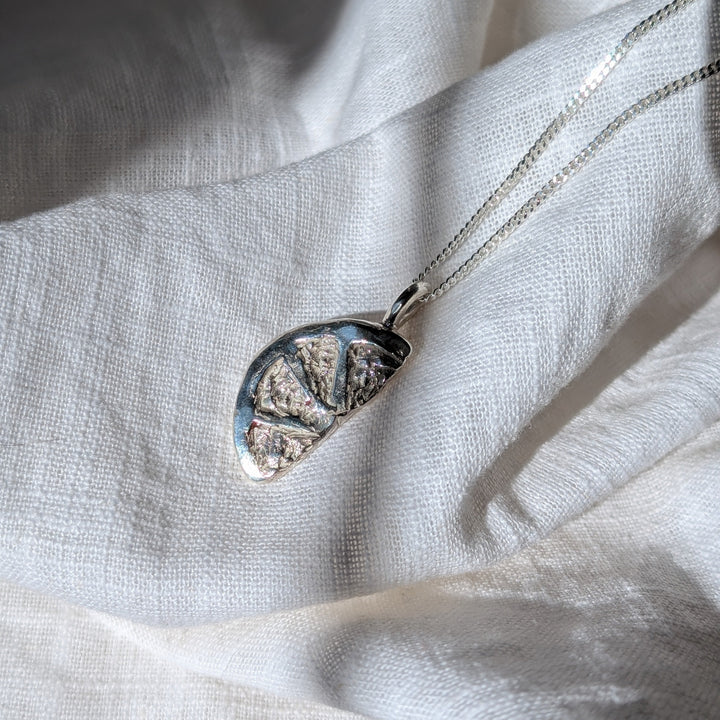 The Becca Calmont Citrus Slice Pendant, a silver necklace with a fruit-slice design and diamond cut curb chain, rests on soft white fabric in natural sunlight, its details shimmering subtly.