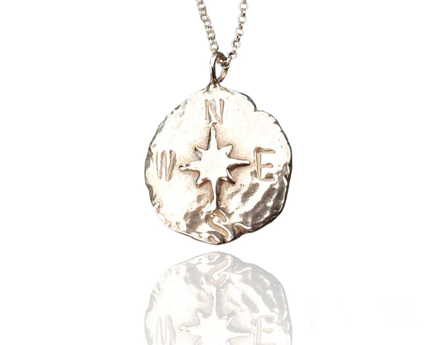 The Pale Raven Compass Coin Silver Necklace features a handmade, irregular sterling silver pendant with a compass rose and "N," "E," "S," "W" letters, crafted in Dublin and finished with a reflective surface.