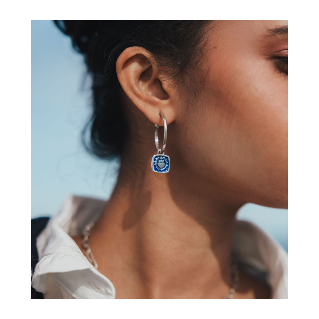 Close-up of a person wearing The Collective Dublin’s Double Sided Keith Haring Claddagh Hoop Earrings. The wearer has smooth skin, their hair tied back, and is styled in a white collared shirt with a silver chain necklace. A blurred blue background accentuates the unique shine of this handmade Dublin jewelry.