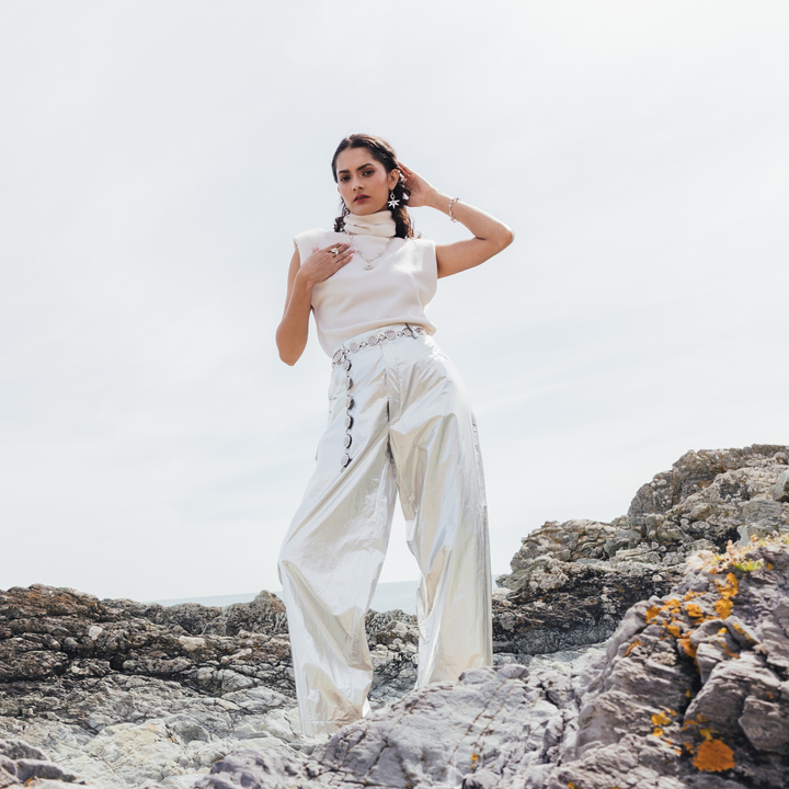 A woman stands on rocky coastal ground wearing a white sleeveless mock-neck top and shiny white wide-leg pants with decorative side details, against a bright overcast sky.
