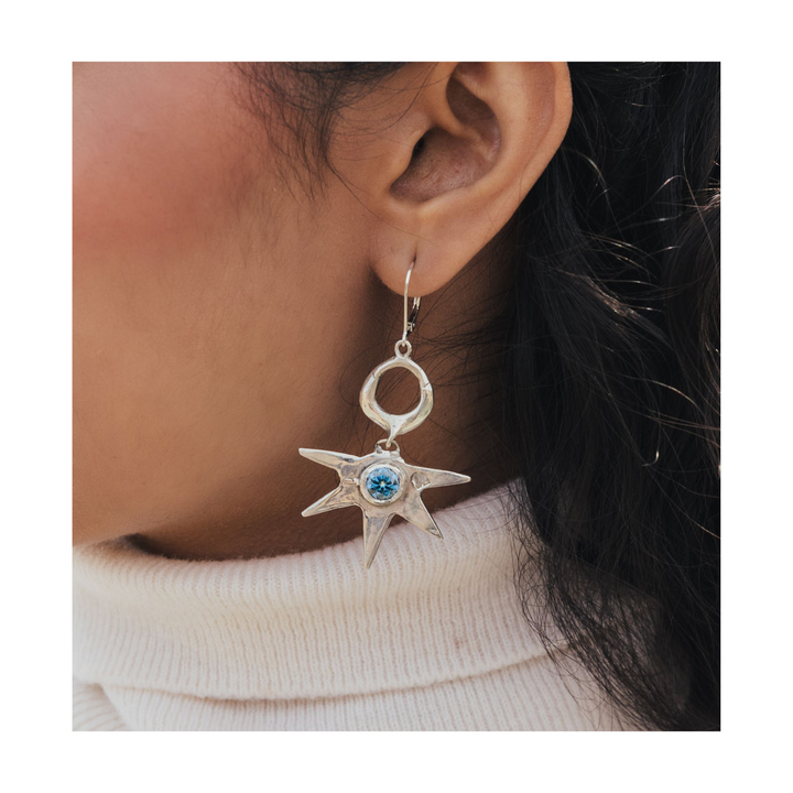 Close-up of a model wearing a textured silver star-shaped drop earring with a round blue gemstone centre.