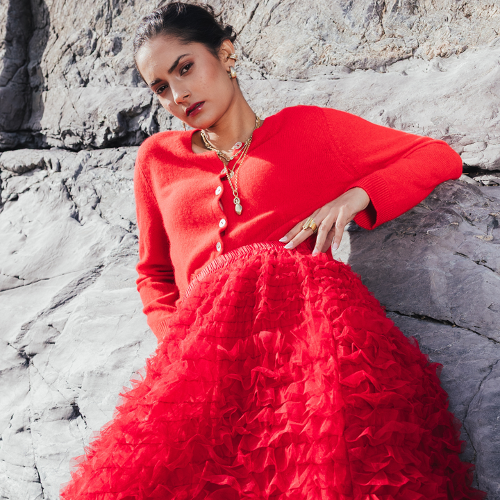 Model wearing the gold-toned Danu Pendant layered with other necklaces over a red cardigan against coastal rocks.