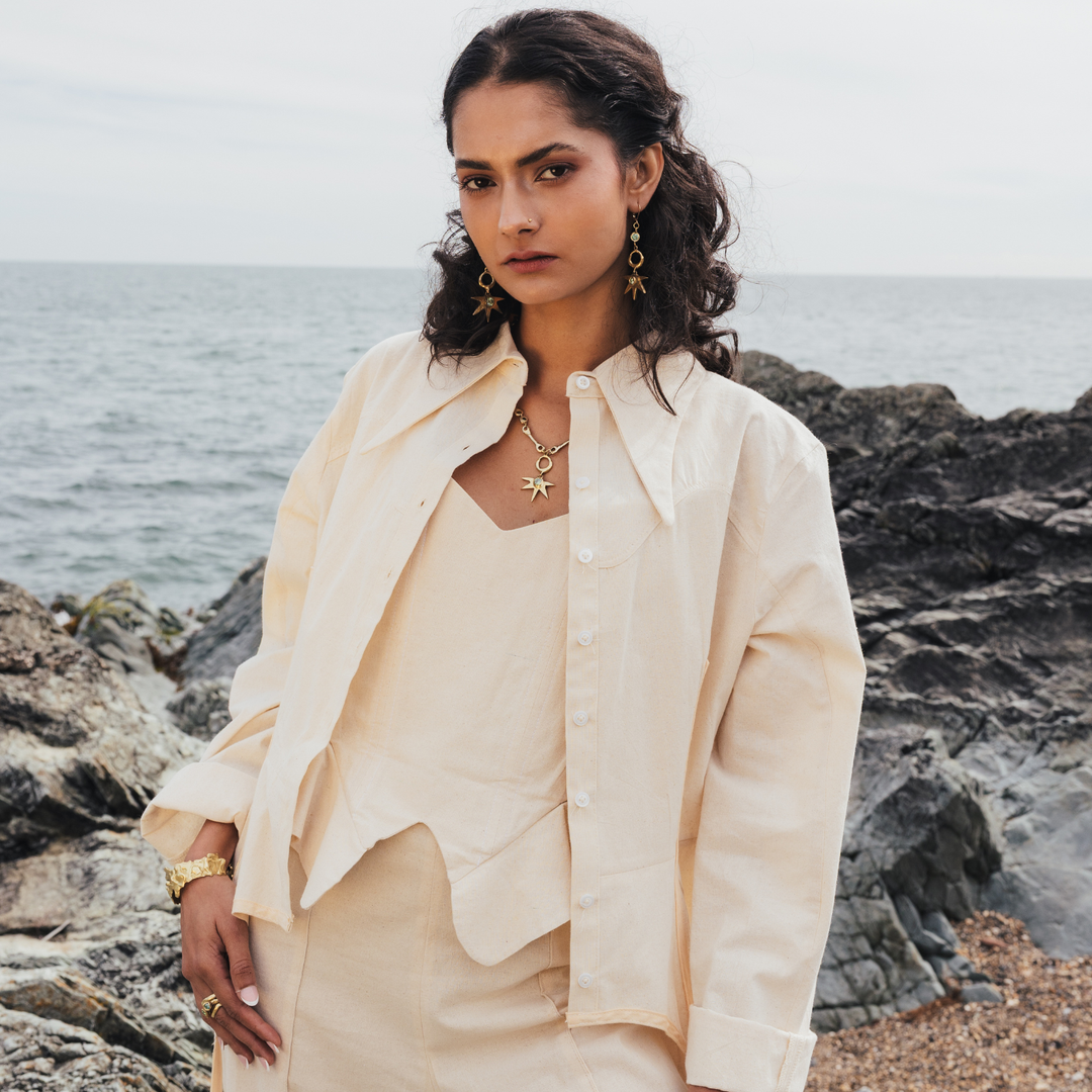 A woman stands on rocky coastal ground with the sea in the background, wearing a cream-colored layered outfit consisting of a button-up overshirt, a sleeveless top, and matching bottoms, accessorized with gold jewelry.