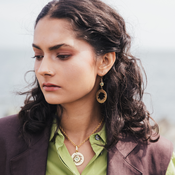 A woman wearing a gold sunburst pendant necklace and matching drop earring, styled with a green shirt and brown blazer against a soft, blurred outdoor background.