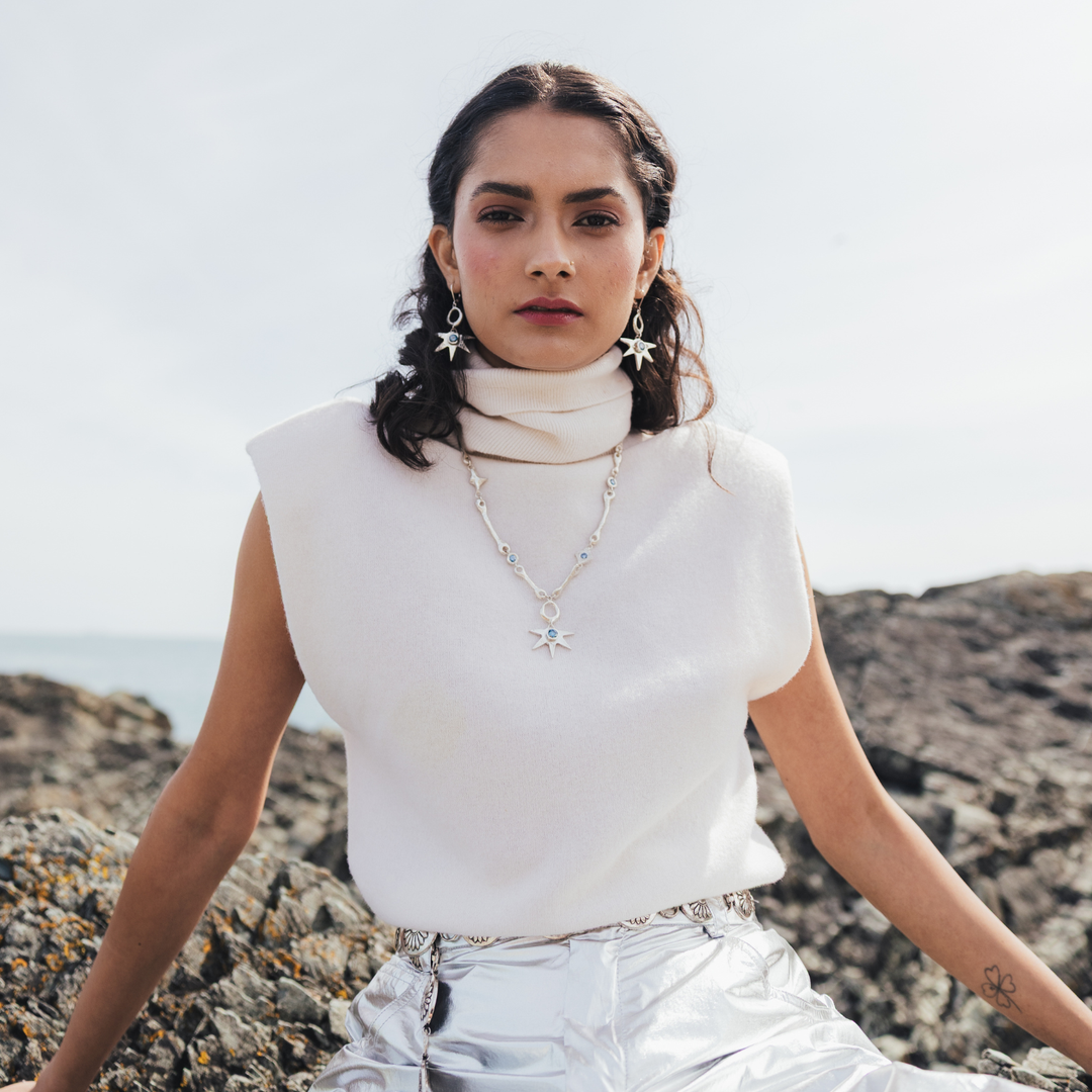 A woman sitting on a rocky beach wearing a white sleeveless mock-neck top, silver metallic pants, and silver star-shaped earrings and necklace.
