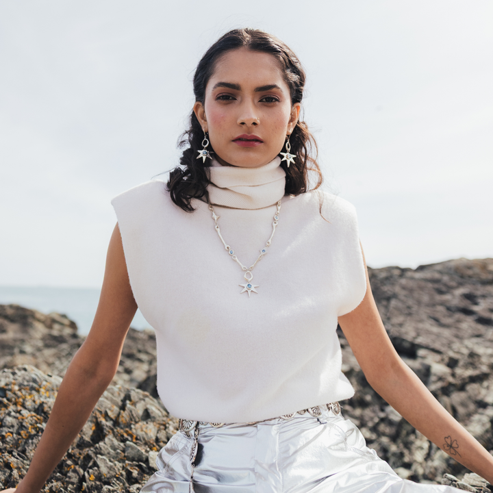 A woman sitting on a rocky beach wearing a white sleeveless mock-neck top, silver metallic pants, and silver star-shaped earrings and necklace.