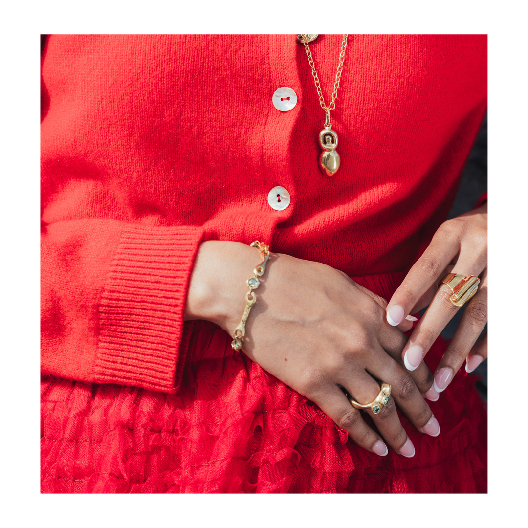 A close-up lifestyle shot of a person wearing gold jewelry, including a delicate gold chain necklace with a double-helix pendant, a gold link bracelet with clear stones, and gold rings, styled with a bright red cardigan and textured red skirt.