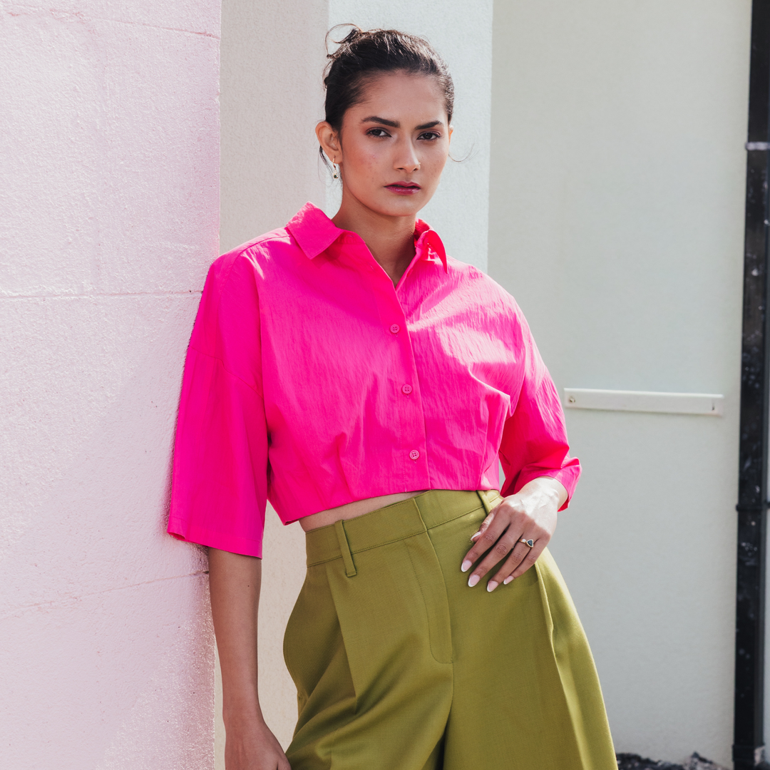 A woman wearing a bright hot pink cropped button-up shirt and olive green trousers, leaning against a light pink textured wall.