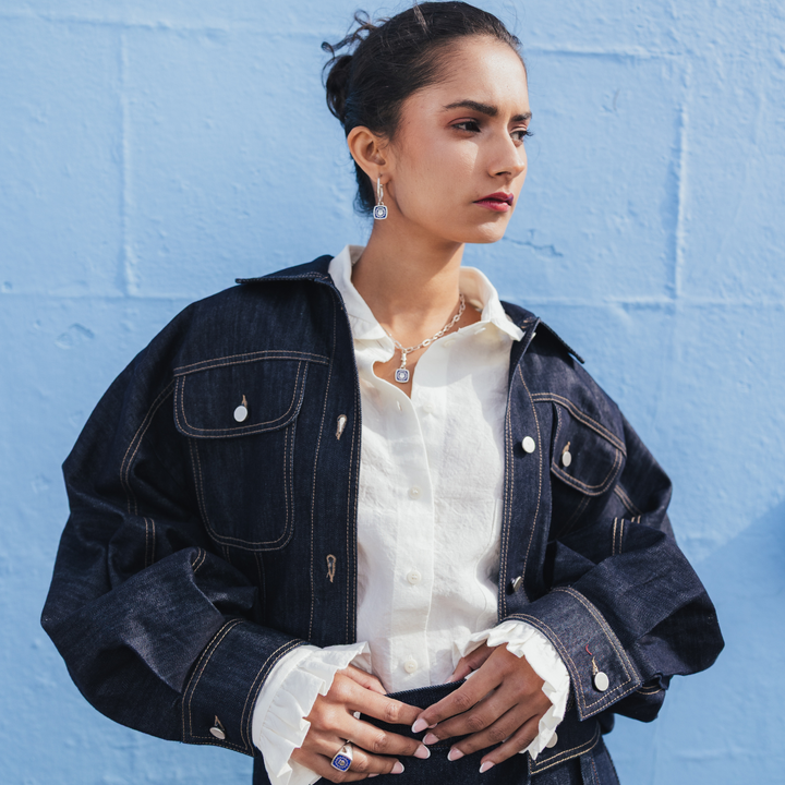 A woman wearing a dark indigo denim jacket over a white ruffled blouse, standing against a light blue painted brick wall.