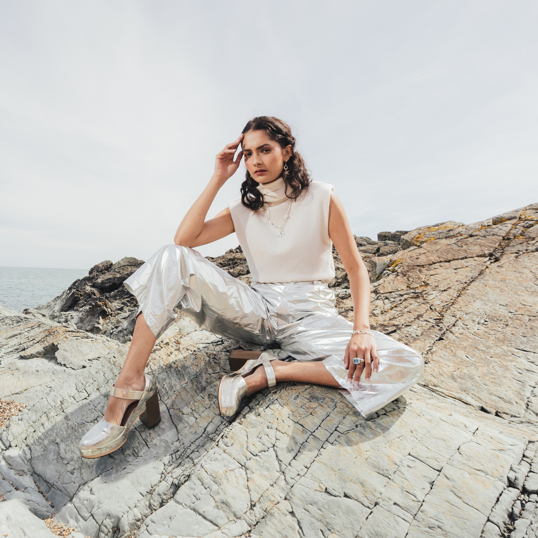 A woman sits on a rocky outcrop by the sea, wearing a sleeveless cream turtleneck top, shiny metallic silver cropped pants, and beige platform sandals.