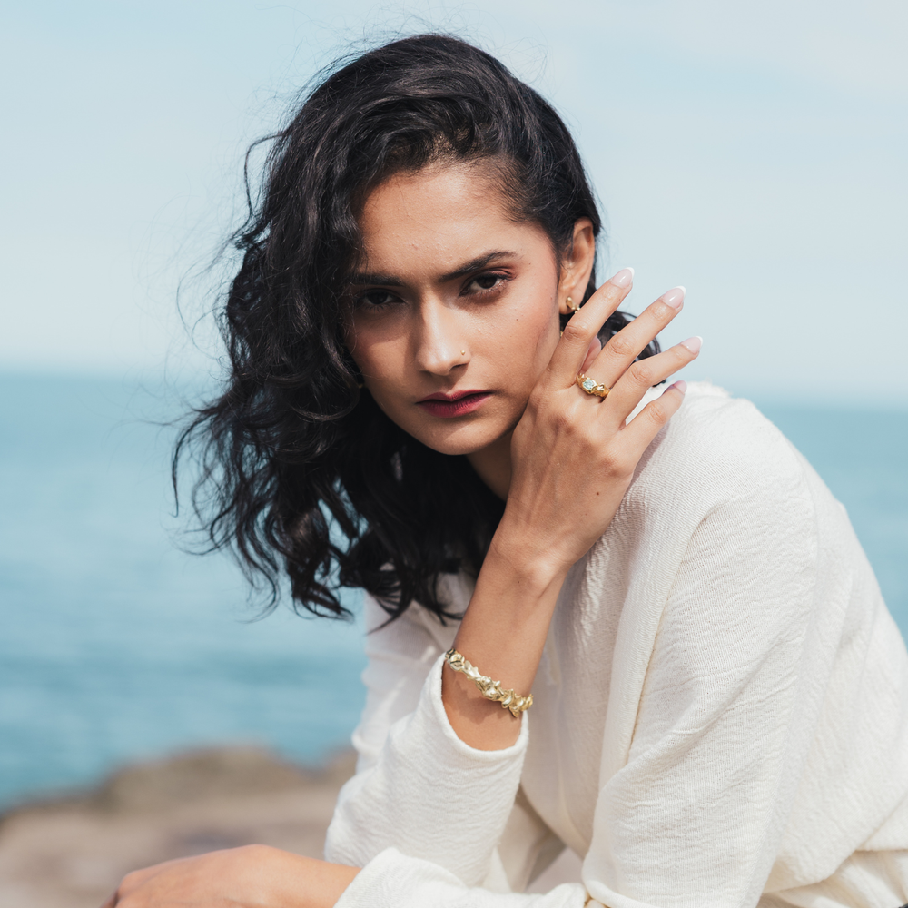 A woman with dark wavy hair wearing a white textured top, gold ring, and gold bracelet, posing outdoors with a soft-focus background of blue sky and sea.
